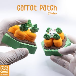 Könnte beinhalten: Zwei "Carrot Patch Clicker" Spielzeuge, jedes mit orangefarbenen Karotten und grünen Blättern in einer grünen und braunen Basis. Eines hat ein weißes Kaninchen. Der Text "Carrot Patch Clicker" ist orange. Die Spielzeuge werden von behandschuhten Händen gehalten.