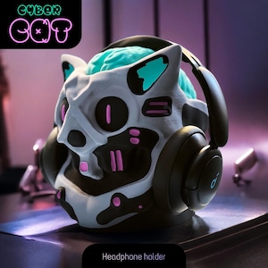 Può includere: Un supporto per cuffie stampato in 3D in bianco e nero a forma di testa di gatto con accenti rosa e blu. Il supporto ha un cervello blu all'interno della testa e il testo "CYBER CAT" in lettere rosa e verde neon sopra il supporto.