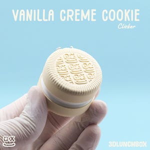 May include: A beige and white cookie-shaped clicker with the text "VANILLA CREME COOKIE" and "Clicker" on a light blue background. The cookie has a textured surface with a repeating design. The clicker is held by a gloved hand.