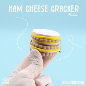 May include: A stack of colorful, miniature ham cheese crackers, held by a gloved hand, against a light blue background. The crackers have pink, yellow, and brown layers. Text at the top reads "HAM CHEESE CRACKER" and "Clicker".