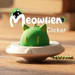 May include: A green cat-shaped clicker toy in a white saucer-like base. The toy has yellow accents and the word "Meowlien" is written above it. The word "Clicker" is written to the right of the toy. A small green beaded chain is in the background.