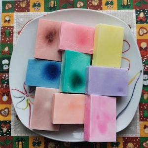 Puede incluir: Ocho barras de jabón artesanal en una variedad de colores, incluyendo rosa, azul, verde, morado y amarillo. Las barras de jabón están dispuestas en una pila sobre un plato blanco.