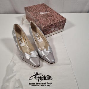 May include: A pair of silver-colored Renata heels with a decorative design on the toe and side. The shoes are displayed with a matching box and a white shoe bag with the Renata logo and text: "Shoes Boots and Bags All Leather - Made in Italy."