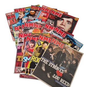 May include: A collection of vintage NME music magazines. The covers feature various artists and bands, with bold red and white mastheads. Some covers have text like "The Strokes" and "Lou Reed". The magazines are stacked on a white background.