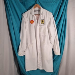 May include: White racing jacket with Shell and Ferrari logos. The jacket has a collared neckline, long sleeves, and a front pocket. The word "FISTIBI" is printed on the collar. The jacket is hanging on a wooden hanger.