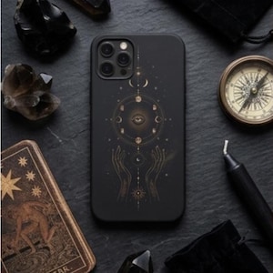 Pode incluir: Capa de telefone preta com um design celestial com luas, um olho e mãos. Rodeada por cristais, uma bússola, cartas de tarô, uma vela preta e pequenas bolsas pretas. A carta de tarô diz "THE STAR."
