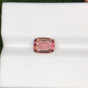 May include: A cushion-cut gemstone with a blend of pink and peach hues. The gem is set against a white background, showcasing its clarity and color. The gemstone's facets reflect light, creating a sparkling effect. This gem is suitable for jewelry.