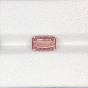 May include: A rectangular, light pink gemstone with rounded corners. The gem has internal lines and facets, giving it a sparkling appearance. The stone is set against a white background, highlighting its color and clarity. This gemstone is suitable for jewelry.