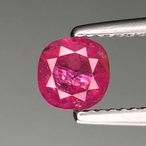 Puede incluir: Un rubí de corte ovalado, sujeto con pinzas. El rubí muestra un tono rojo intenso con un patrón en forma de estrella en el centro. La gema se encuentra sobre un fondo gris neutro, resaltando su color y claridad.