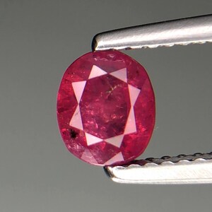 Puede incluir: Una gema de rubí facetada, de forma ovalada, sujeta con pinzas. El rubí es de color rojo intenso con un patrón en forma de estrella de seis puntas visible en su interior. El fondo es gris neutro.