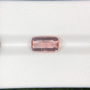 May include: A rectangular, light pink gemstone with rounded corners. The gem has internal lines and facets, giving it a polished appearance. The stone is set against a white background, highlighting its color and clarity.