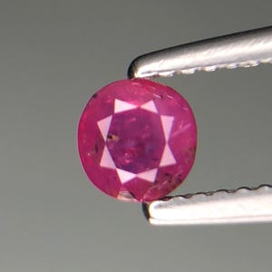 Puede incluir: Una gema de rubí redonda y facetada sujeta con pinzas. El rubí es de color rojo rosado intenso con un patrón en forma de estrella de facetas blancas en el centro. Las pinzas son de color plateado.
