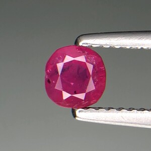 Puede incluir: Una gema de rubí facetada, de forma ovalada, sujeta con unas pinzas plateadas. El rubí es de un rojo intenso y vibrante, con un patrón en forma de estrella visible en el centro. Se muestra sobre un fondo gris neutro.