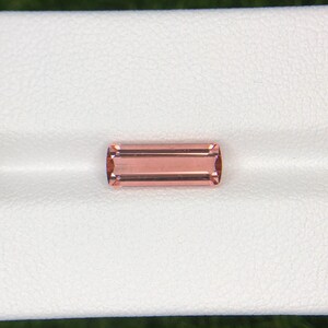 May include: A rectangular, peach-colored gemstone with a polished surface. The gem has a subtle, warm hue and is set against a white background. The gemstone's shape is a long rectangle with slightly rounded corners, showcasing its clarity and color.