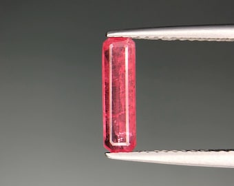 May include: A vibrant, rectangular, pink gemstone is held by tweezers against a gray background. The gemstone has a polished, transparent appearance, with visible internal structures. The tweezers are silver-colored, and the overall composition highlights the gem's color and form.