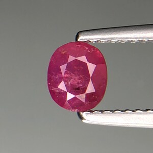 Puede incluir: Una gema de rubí facetada, de forma ovalada, sujeta por unas pinzas plateadas. El rubí es de un rojo intenso y vibrante, con facetas internas que crean un patrón de estrella. La gema se encuentra sobre un fondo gris neutro.