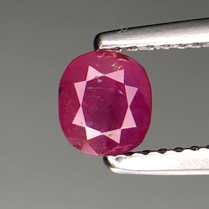 Puede incluir: Un rubí facetado de forma ovalada, sostenido por unas pinzas. El rubí es de color rojo intenso y presenta un patrón en forma de estrella en su interior. Las pinzas son plateadas y el fondo es gris neutro. Medidas en milímetros.