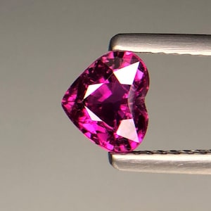 May include: A heart-shaped gemstone with a vibrant magenta hue and a faceted surface. The stone is held by a pair of metallic tweezers, showcasing its clear, reflective quality and precise cut. The gem displays deep pink tones and sharp edges.