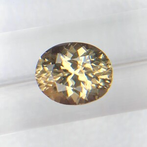 Puede incluir: Una gema de corte ovalado con una superficie facetada. La gema tiene un color marrón dorado cálido y refleja la luz, creando un efecto brillante. La gema se encuentra sobre un fondo blanco, resaltando su claridad y brillo.