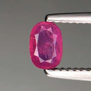 Puede incluir: Una gema de rubí facetada, de forma ovalada, sujeta con unas pinzas plateadas. El rubí es de un color rojo-rosado intenso y vibrante, con facetas internas que crean un patrón en forma de estrella. El fondo es gris neutro.