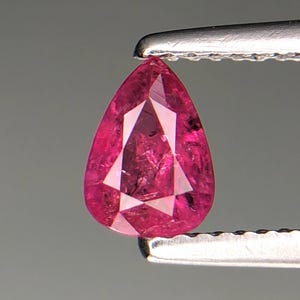 Puede incluir: Una gema facetada en forma de pera, de color rosa intenso, sujeta con unas pinzas plateadas. La gema tiene una estructura interna compleja con inclusiones visibles y un alto grado de transparencia. La gema es de un tono rosa vibrante.