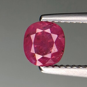 Puede incluir: Gema de rubí de corte ovalado sujeta con pinzas. El rubí es de color rojo intenso con facetas que crean un patrón de estrella en el centro. La gema está sobre un fondo gris.