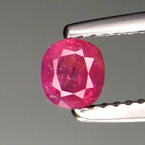 Puede incluir: Gema de rubí ovalada y facetada, sujeta con pinzas. El rubí es de un color rojo rosado intenso, con un patrón en forma de estrella visible en su interior. Las pinzas son plateadas y el fondo es gris neutro.