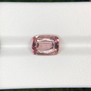 May include: A cushion-cut gemstone with a soft pink hue and subtle internal lines. The gem has a rectangular shape with rounded corners, and it is set against a white background. The stone's facets reflect light, creating a sparkling effect.