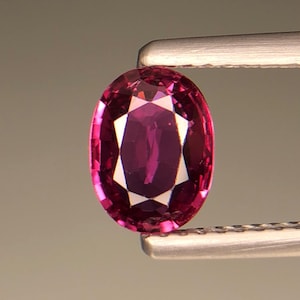 May include: An oval-cut gemstone with a deep magenta and purple hue, featuring a faceted surface that creates a symmetrical pattern of light and dark reflections. The stone is held by a pair of metallic tweezers with a textured grip.