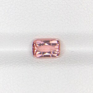 May include: A faceted, rectangular-cut gemstone with a soft pink hue. The gem has a polished surface with multiple facets that reflect light. The gemstone is set against a white background, highlighting its color and clarity. This gem is suitable for jewelry making.