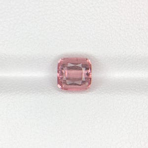 May include: A square-cut, light pink gemstone with a faceted surface. The gem is set against a white background, showcasing its clarity and color. The gemstone's shape is a perfect square, with a subtle, internal pattern.