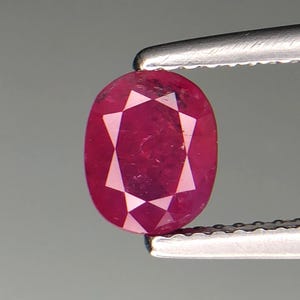 Puede incluir: Una gema de rubí ovalada y facetada sujeta con pinzas. El rubí es de color rojo intenso con facetas internas que crean un patrón de estrella. Las pinzas son plateadas y el fondo es gris neutro.