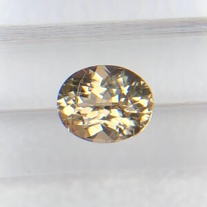 Puede incluir: Una gema ovalada y facetada, de color marrón claro. El tallado brillante de la gema refleja la luz, creando un efecto brillante. La gema está sobre un fondo blanco. Mide aproximadamente 1,5 cm de largo.