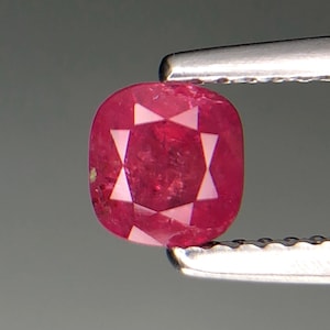 Puede incluir: Una gema de rubí facetada, de talla cojín, sujeta con pinzas. El rubí es de color rojo intenso con un patrón en forma de estrella de facetas blancas. La gema mide aproximadamente 1 cm de largo.