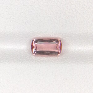 May include: A rectangular, light pink gemstone with rounded corners. The gem has a polished surface and internal facets that reflect light. The stone is set against a white background, highlighting its color and clarity. This gemstone could be used in jewelry.