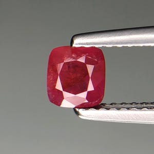 Puede incluir: Un rubí de talla cojín de color rojo intenso, sujeto con pinzas. Las facetas del rubí reflejan la luz, creando un patrón en forma de estrella. La gema es cuadrada con esquinas redondeadas.