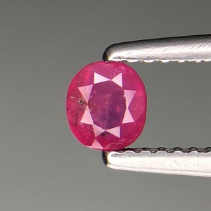 Puede incluir: Una gema de rubí facetada, de forma ovalada, sujeta con unas pinzas plateadas. El rubí es de un rojo intenso y vibrante con facetas internas en forma de estrella. El fondo es gris neutro.