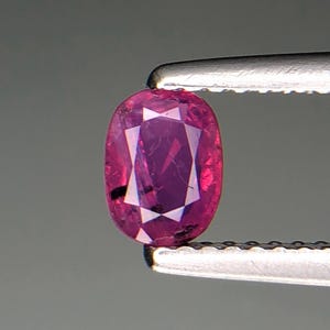 Puede incluir: Una gema de rubí facetada, de forma ovalada, sujeta con unas pinzas. El rubí es de color rojo rosado intenso con inclusiones internas. La gema mide aproximadamente 1,3 cm de largo.
