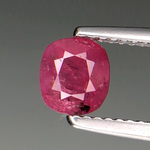 Puede incluir: Una gema de rubí facetada, de forma ovalada, sujeta con unas pinzas plateadas. El rubí es de color rojo rosado intenso con un patrón en forma de estrella en el centro. La gema tiene una superficie pulida sobre un fondo gris.