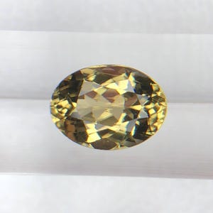 Puede incluir: Una gema de corte ovalado con una superficie facetada. La gema tiene un color amarillo verdoso y está colocada sobre un fondo blanco. Las facetas de la gema reflejan la luz, creando un efecto brillante.