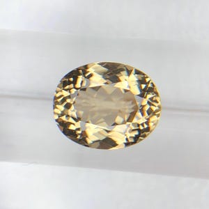 Puede incluir: Gema de corte ovalado con superficie facetada. La gema tiene un color marrón dorado cálido y refleja la luz, creando un efecto brillante. La gema se encuentra sobre un fondo blanco, resaltando su claridad y brillo.