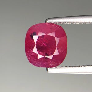 May include: A faceted, oval-shaped ruby gemstone, held by tweezers. The ruby displays a deep red hue with a star-like pattern in the center. The gemstone is set against a neutral grey background.