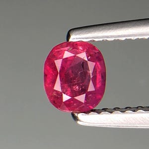 Puede incluir: Una gema de rubí facetada, de corte ovalado, sujeta con pinzas. El rubí es de un color rojo intenso y vibrante con facetas internas que reflejan la luz. Las pinzas son de color plateado y el fondo es gris neutro.