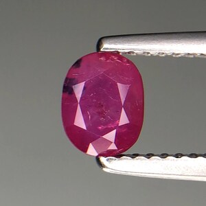 May include: A faceted, oval-shaped ruby gemstone held by silver tweezers. The ruby displays a deep red hue with internal facets creating a starburst pattern. The gemstone is set against a neutral grey background.