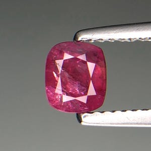 Puede incluir: Una gema de rubí tallada en cojín, sujeta con pinzas plateadas. El rubí es de color rojo intenso con un patrón en forma de estrella en el centro. La gema mide aproximadamente 1,3 cm de largo.