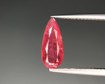 Puede incluir: Una gema facetada en forma de lágrima sujeta con pinzas. La gema es de color rojo vibrante con facetas internas que reflejan la luz. El fondo es gris sólido y apagado.