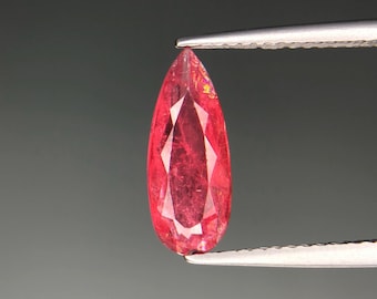 Puede incluir: Una gema facetada en forma de lágrima sostenida por unas pinzas. La gema es de color rojo vibrante con facetas internas visibles. El fondo es gris neutro.