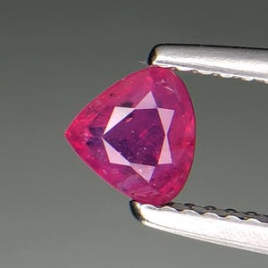 Puede incluir: Una gema de rubí facetada, de forma triangular, sujeta con pinzas. El rubí es de un color rojo rosado vibrante con un patrón en forma de estrella visible en el centro. La gema está tallada con múltiples facetas para reflejar la luz.