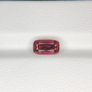 May include: A cushion-cut, reddish-pink gemstone with facets. The gem is rectangular with rounded corners and is set against a white background. The gemstone's color is a deep, saturated pink.