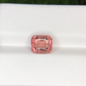May include: A cushion-cut, peach-colored gemstone. The gem is set against a white background. The gemstone has a square shape with rounded corners and internal facets that reflect light.
